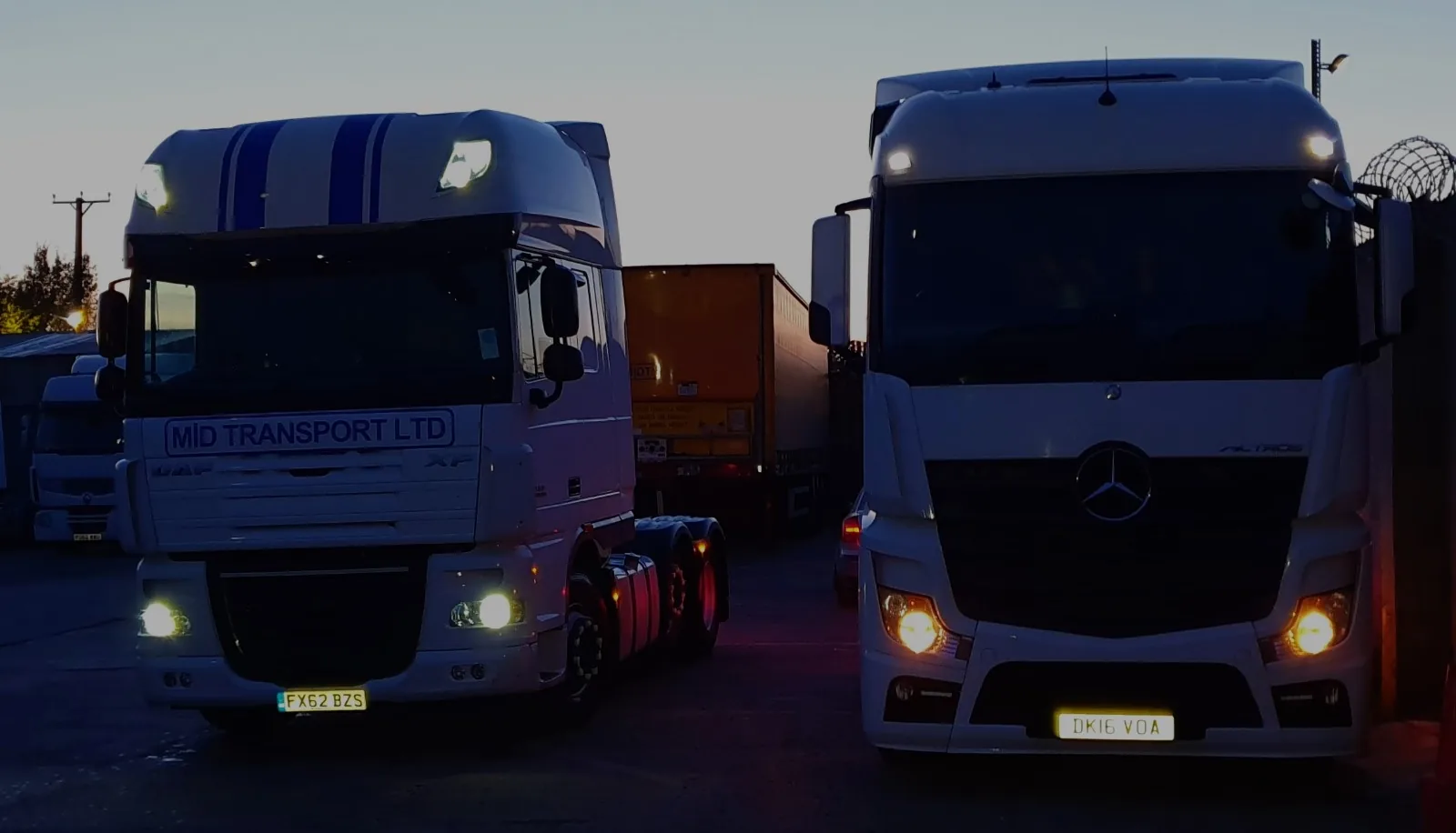 Two Mid Transport tractor units — a liveried DAF XF and a Mercedes-Benz Actros — with a curtainside trailer in the yard at dusk.